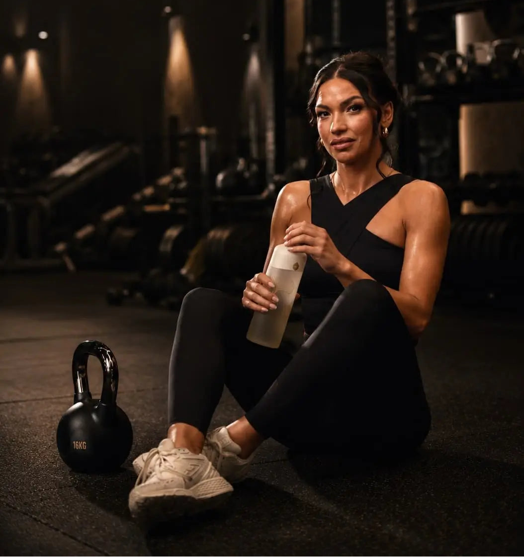 Woman sitting on the floor in a gym holding a bottle, with a kettlebell nearby.
